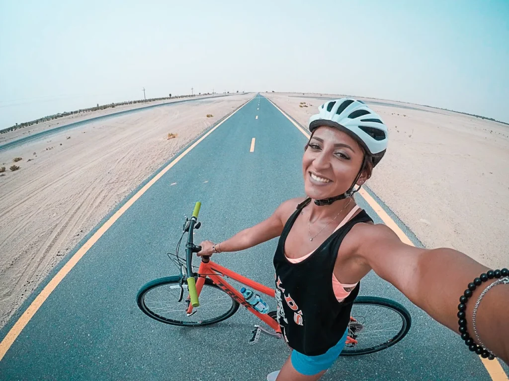 Smiling woman taking a selfie while cycling on a long, open desert road in Dubai, wearing a helmet and athletic gear with clear skies above.