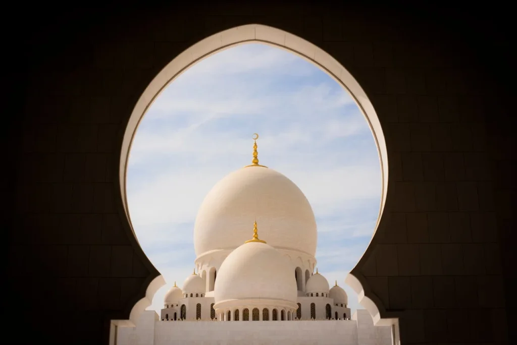 View of the domes of Sheikh Zayed Grand Mosque in Abu Dhabi, framed by an ornate arched window, with a clear blue sky in the background.