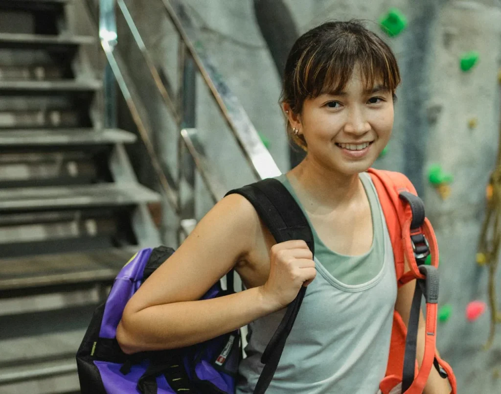 Smiling young woman in athletic wear carrying climbing gear and a backpack, standing indoors near a staircase and a rock climbing wall.