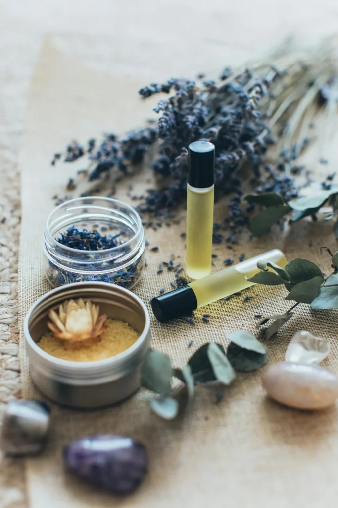 Small cork-topped glass bottles of massage oil with a sprig of lavender and soft purple flowers on a natural woven surface
