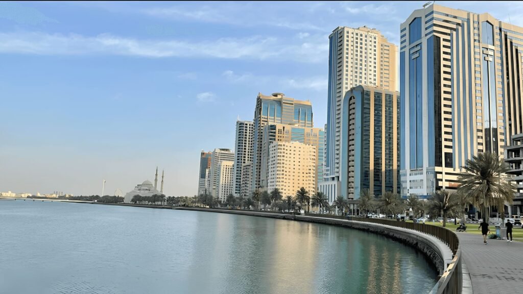 Scenic view of Sharjah Corniche Beach with modern high-rise buildings, calm waterfront promenade, and clear blue sky